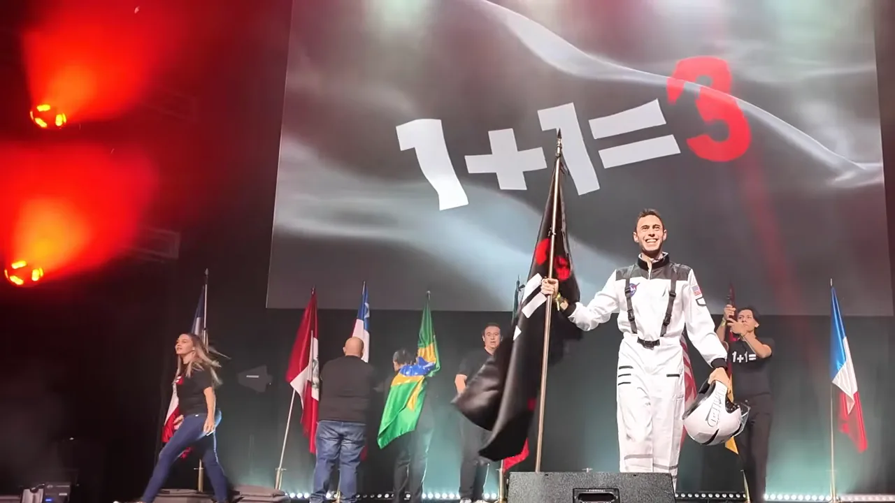 Manuel de León en el escenario con traje blanco y la bandera «1+1=3», frente a una pantalla con el mismo lema