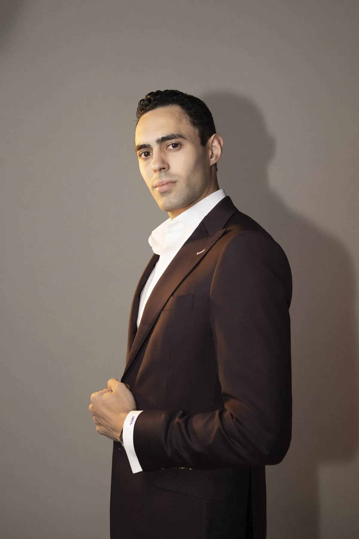 Manuel de León de pie, con saco café y camisa blanca, mira a la cámara en un estudio de fondo gris